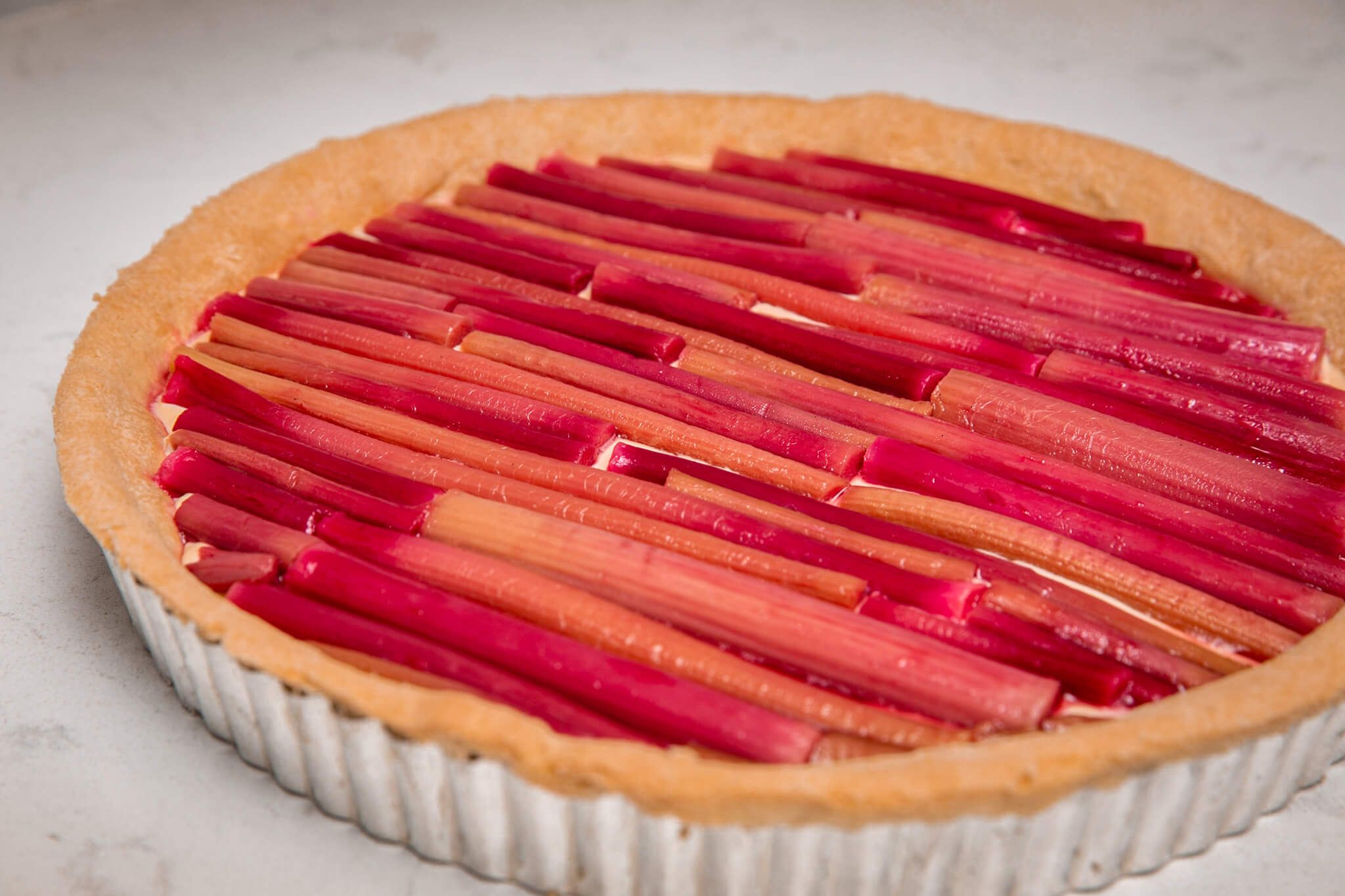 A large tart with pastry cream and topped with rows of rhubarb stems