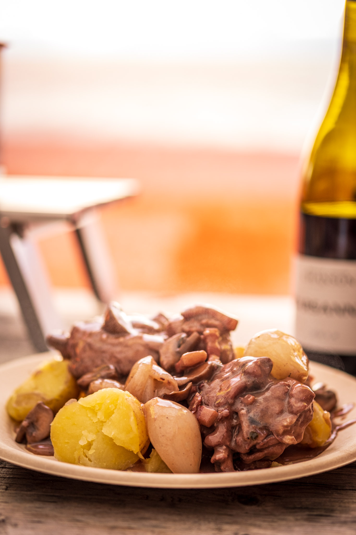 Plate of coq au vin with potatoes and a bottle of Burgundy wine on a campground table