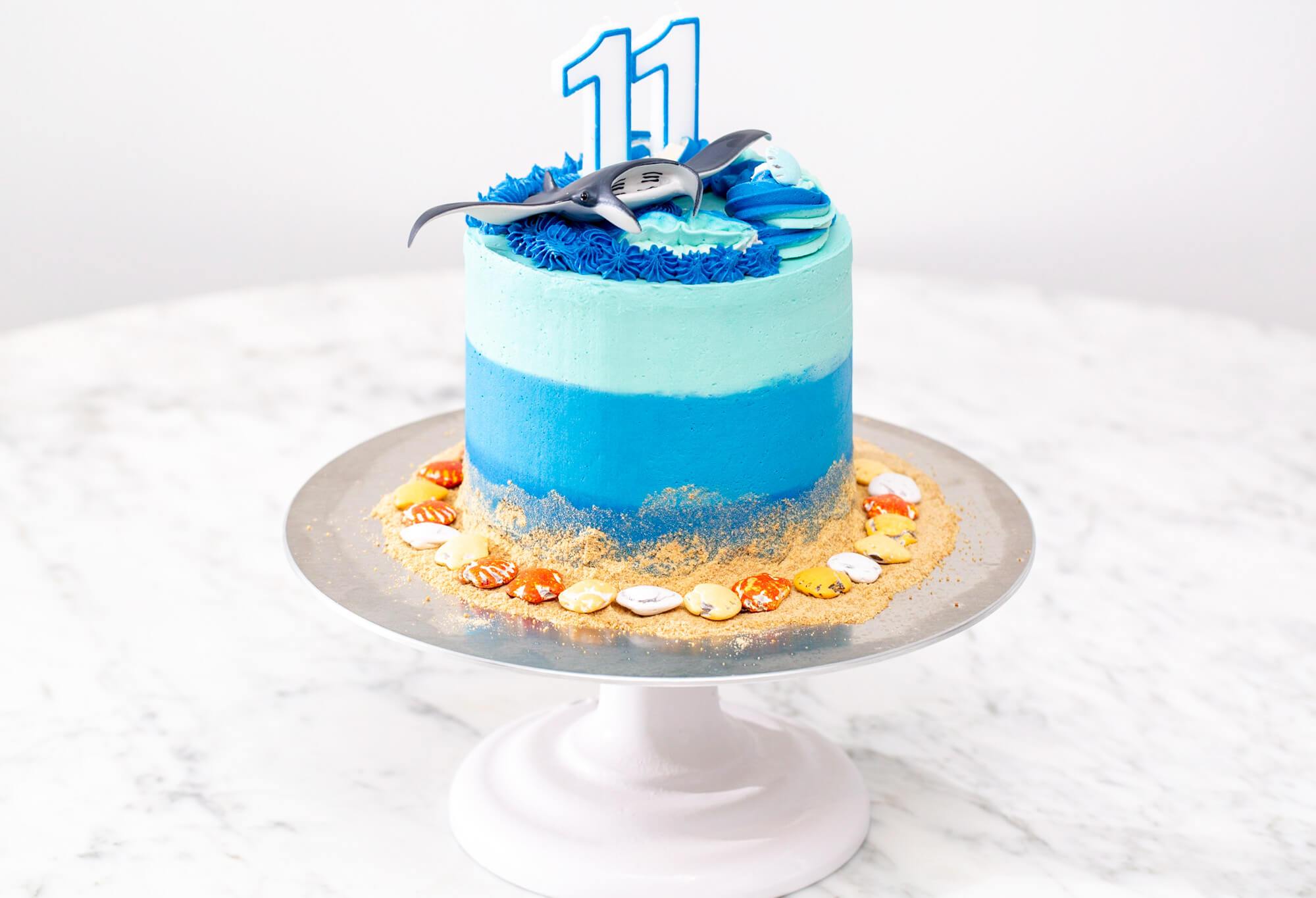A blue cake decorated with an underwater theme featuring chocolate shells, graham crumb sand and a toy manta ray